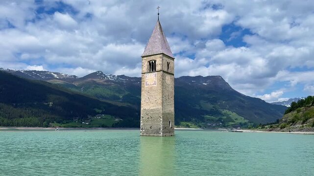 Extended steady view of the submerged Curon bell tower at Lake Resia with rippling water, green slopes and snow-dusted Alpine peaks.