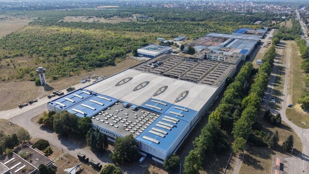 Aerial view of the Severna Zona industrial complex featuring large factory buildings with blue roofs, surrounding green fields, and access roads in Pancevo, Serbia.