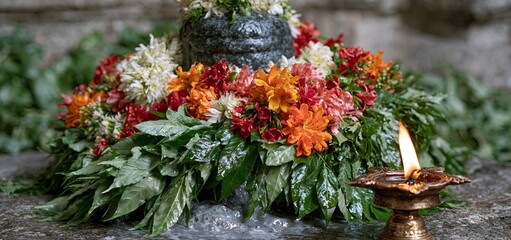 Fototapeta premium A sacred Shiva Lingam adorned with lush leaves and colorful flowers, illuminated by an oil lamp in a peaceful temple.