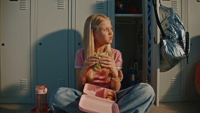 Blonde Elementary School Girl Enjoys Fresh Sandwich in Pink Reusable Lunch Box While Sitting Beside Metal Lockers in Bright School Hallway. Nutrition Habits, Childhood Independence, Modern Education.