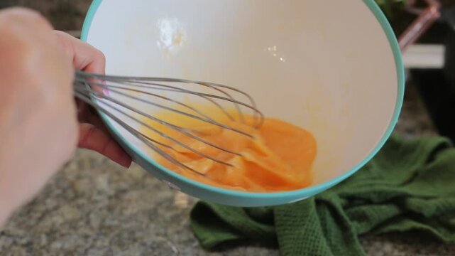 A hand whisks raw eggs with a metal whisk inside a blue polka-dot bowl, blending the yolks into a smooth mixture for homemade baking