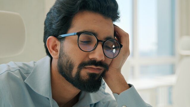 Tired Indian business man sleepy exhausted office worker at workplace bored Arabian entrepreneur leaning hand laptop computer low energy burnout fatigue stress depression job routine exhaustion indoor