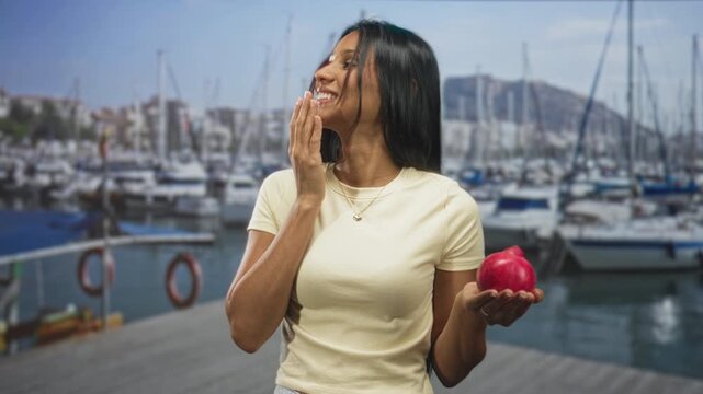 Woman holding pomegranate with hand to mouth gesture on a marina dock by boats and yachts on a street; playful delight.