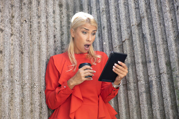 Young woman in red shirt reacting with surprise and shock while reading news on a tablet and holding a coffee cup outdoors © Dejan