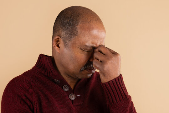 Mature African American man pinching the bridge of his nose with closed eyes on a beige studio background, representing stress, fatigue, sinus pain, headache and emotional exhaustion