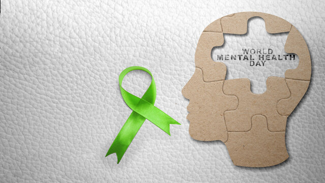 Green ribbon next to a head-shaped cardboard puzzle with a missing piece showing World Mental Health Day text on textured background. Concept of World Mental Health Day