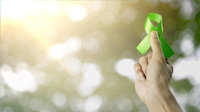 Hand holding a green awareness ribbon against a sunny, blurred nature background. Symbol for mental health, depression, organ donation, and environmental causes. Concept of World Mental Health Day
