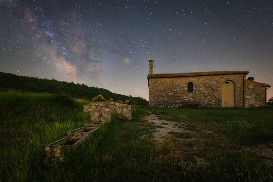 View of the historic stone church of Madonna Aguzza under a starry night sky with the Milky Way visible above a grassy field and stone water trough in Fiordimonte, Marche, Italy.