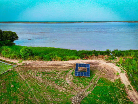 Aerial view of solar panels installed on the bank of the Indus River surrounded by lush green fields and dirt paths in Dera Ghazi Khan, Punjab, Pakistan.