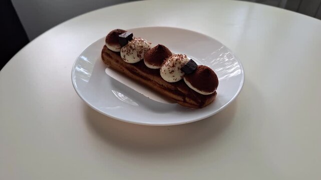 A close-up of an exquisite chocolate eclair covered with swirls of cream and pieces of chocolate is depicted on a white dish