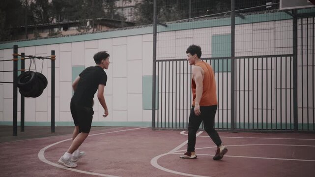 Two young men play basketball on an outdoor court in an urban setting, dribbling and smiling during a friendly game