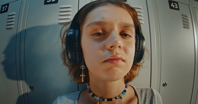 Close Up Portrait of Curly Haired Teenage Girl Wearing Headphones and Listening to Music in High School Hallway. Relaxed and Thoughtful Expression, Direct Gaze, Youth Lifestyle and Gen Z Identity.