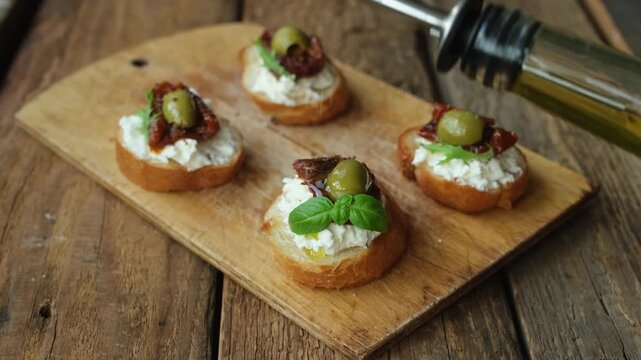 Cooking bruschetta or crostini. Pouring olive oil over a toasted bread with cream cheese, sun dried tomatoes and olives.