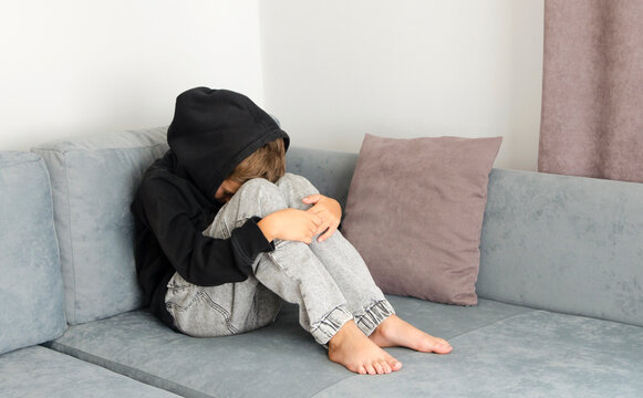 Boy crying with his head down and sitting sad scared on the sofa in his room and hugs his knees. Concept domestic violence and child abuse.