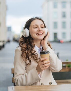 Cheerful girl wearing white headphones enjoying audio content at sidewalk terrace, happy young woman with coffee cup smiling with closed eyes 