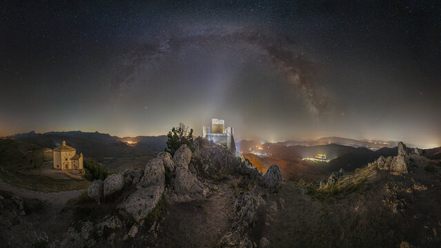 View of Rocca Calascio illuminated under a starry night sky with the Milky Way arching over the historic fortress and nearby church in the mountains. Calascio, Abruzzo, Italy.