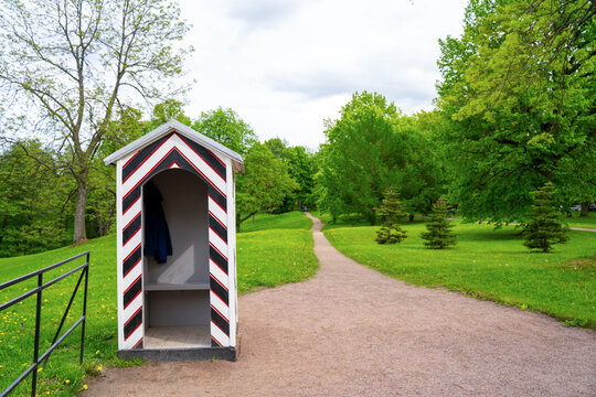 A striped black white and red sentry box stands beside a winding gravel path in a lush park with dandelions and tall trees, a historic landmark and quiet outdoor retreat.