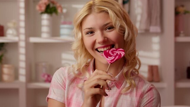 Laughing glad Caucasian teen girl biting heart shaped lollipop in pink bathroom looking at camera playful female woman enjoying sugary candy delight sweet tooth mood glamour beauty fun home interior