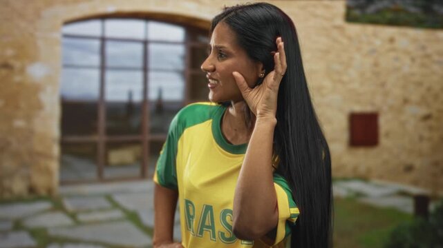 Woman cupping ear and grimacing while wearing yellow brazil t shirt in a stone building; concern listening.