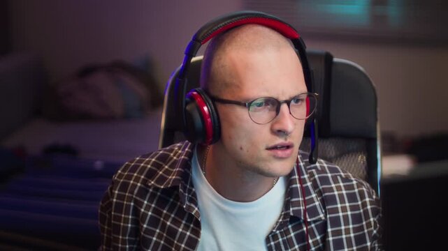 Young adult Caucasian man wearing headphones and glasses staring intensely at computer screen, playing video games in slow arc shot. Footage represents screen addiction or esports gaming concepts