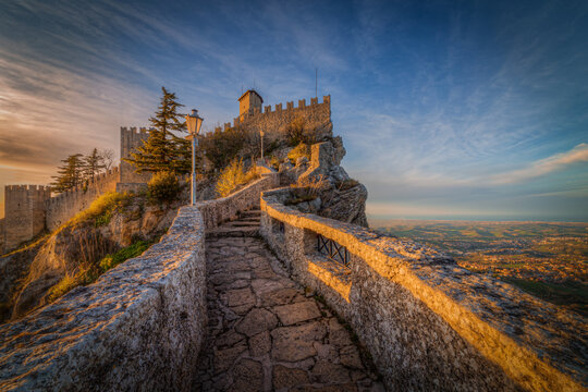 View of the historic Guaita Tower and stone walkway atop Mount Titano at sunset with panoramic views of the surrounding landscape in San Marino, Emilia-Romagna, Italy.