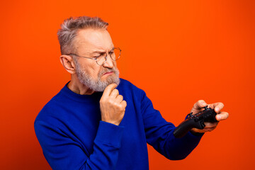 Elderly man concentrating with video game controller, puzzled thoughtful expression while gaming against bright orange background © deagreez