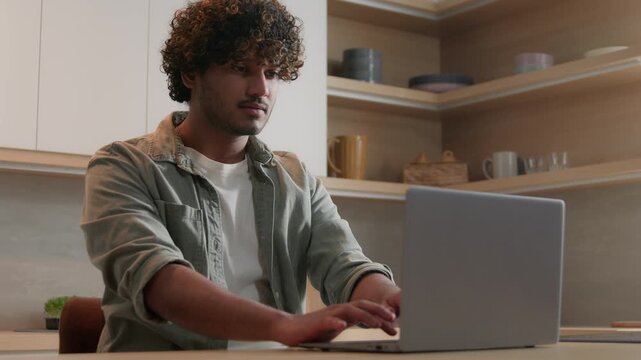 Indian Arabian business man working on laptop at home kitchen focused typing computer remote work ethnic male freelancer shopping online study e-learning serious businessman task concentration indoor