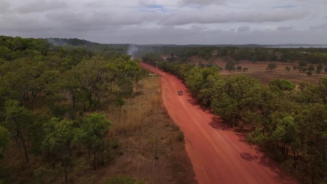 Aerial view of vehicle travelling along a dusty red dirt road in remote outback Australia, with a small grass fire burning beside the road. Regional transport, bushfire risk, remote landscape concept
