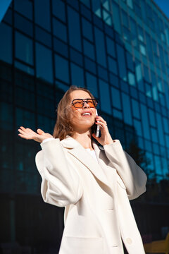 Happy businesswoman talking on smartphone with expressive gesture outdoors. Professional woman enjoying phone conversation outside office building, communication