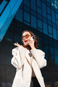 Happy businesswoman talking on smartphone with cheerful smiling expression. Professional woman enjoys successful phone conversation outside office building, business communication =