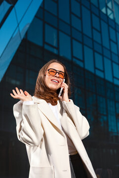 Cheerful businesswoman talking on smartphone with joyful confident smile. Professional woman enjoying phone conversation beside modern office building, 