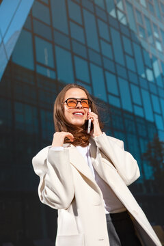 Smiling businesswoman talking on smartphone with relaxed happy expression. Professional woman enjoying successful phone conversation outside office building, 