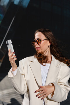 Professional businesswoman looking at smartphone with surprised curious expression. Confident woman checking mobile phone beside modern office building, 