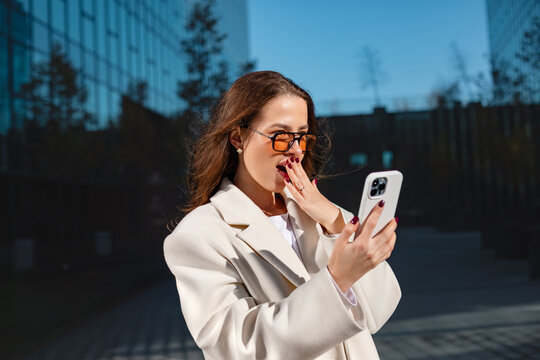 Surprised businesswoman looking at smartphone with playful smiling expression. Professional woman reacts to mobile phone beside modern office building, digital communication 