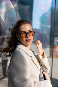 Portrait of female model adjusting sunglasses with elegant confident style. Businesswoman holding smartphone beside modern office building, professional fashion and corporate lifestyle concept.