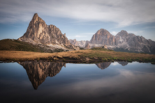 View of the dramatic peaks of Passo Giau reflecting in a calm alpine pond surrounded by golden grasses and rocky mountain terrain in Colle Santa Lucia, Veneto, Italy.