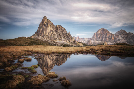 View of Passo Giau with its dramatic mountain peaks reflecting in a calm alpine pond under a cloudy sky in Colle Santa Lucia, Veneto, Italy.