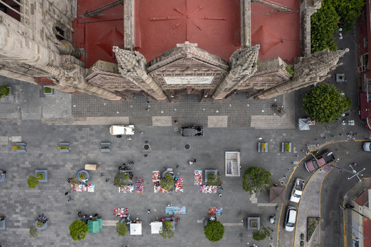 Aerial view of Parroquia El Expiatorio Eucaristico with its neo-Gothic facade and red roof, overlooking a plaza with street vendors and parked cars in Guadalajara, Jalisco, Mexico.