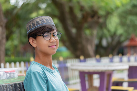 Portrait of a smiling Muslim boy wearing a traditional cap and eyeglasses, sitting outdoors in a park with a blurred green background.