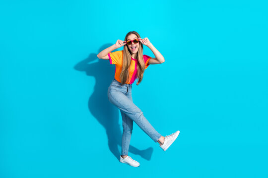 Young woman in bright shirt and jeans jumps with sunglasses against a blue background