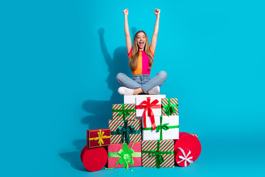 Joyful teen girl on colorful gifts celebrating with arms up in bright blue studio