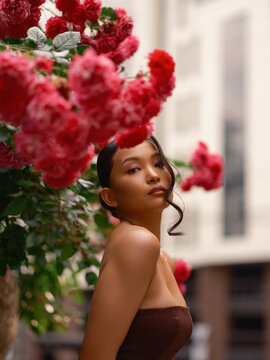 Asian Woman Looking At Camera Near Blooming Pink Roses Outdoors