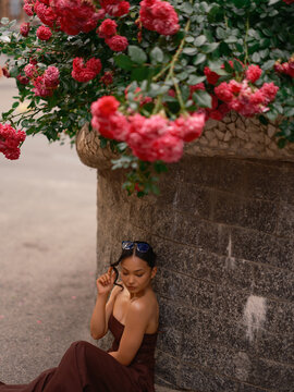 Asian Woman Sitting Near Stone Wall Under Blooming Roses