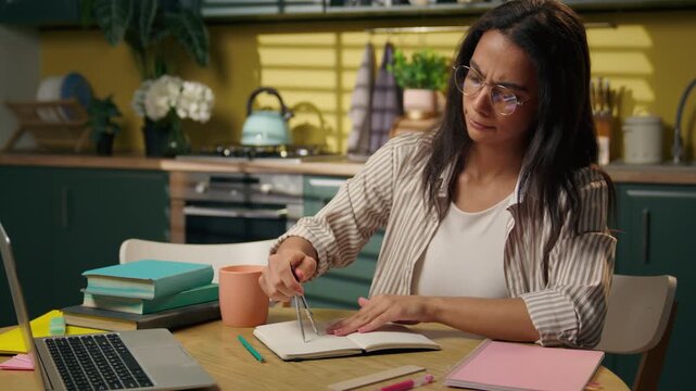 Focused Indian Arabian Latina teen woman studying at home writing notes with compass student pupil freelancer drawing draft in notebook pencil university high school geometry distant study at kitchen