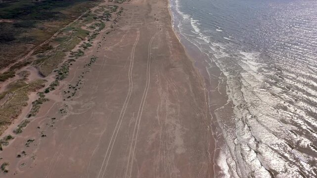 Aerial view video clip of Katwijk aan Zee sandy beach, tire tracks on sand, coastal dunes landscape, sunlight reflection on waves, and North Sea horizon in South Holland, Netherlands