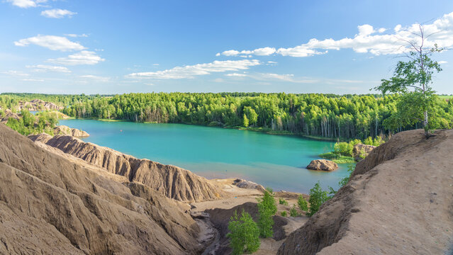 Scenic view of turquoise lake surrounded by lush green forest and clay hills under a clear blue sky with scattered clouds in a natural landscape in Konduki, Tula region, Russia
