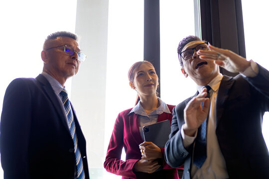 Three businesspeople discussing strategy by an office window, with one man gesturing while colleagues listen, showing leadership, teamwork, planning, communication, and professional collaboration
