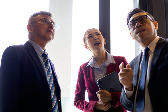 Three businesspeople standing by an office window looking upward while discussing strategy, teamwork, leadership, communication, planning, collaboration, management, and professional ideas