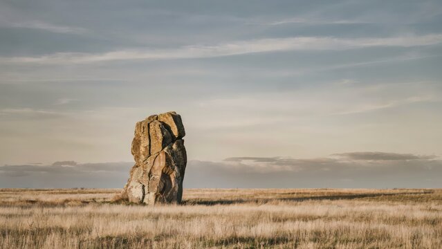 A solitary large weathered stone monolith standing in the middle of a vast dry grassy field under a soft overcast sky during the late afternoon