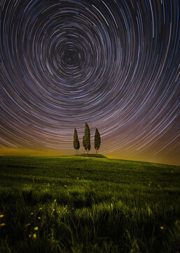 View of the iconic cypress trees of San Quirico d'Orcia under a dramatic star trail night sky in the rolling hills of the Val d'Orcia, San Quirico d'Orcia, Tuscany, Italy.